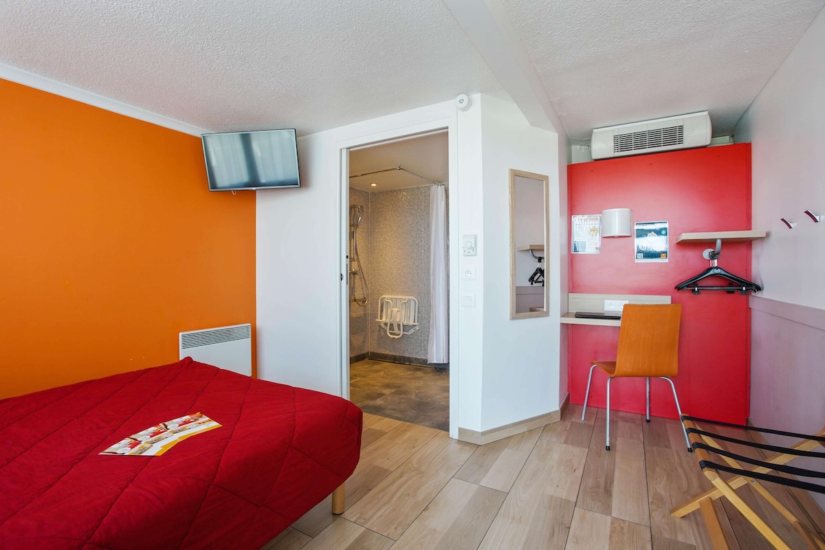 Een kamer met een rode bed, een tv aan de muur, een rode stoel en een klein keukentje.