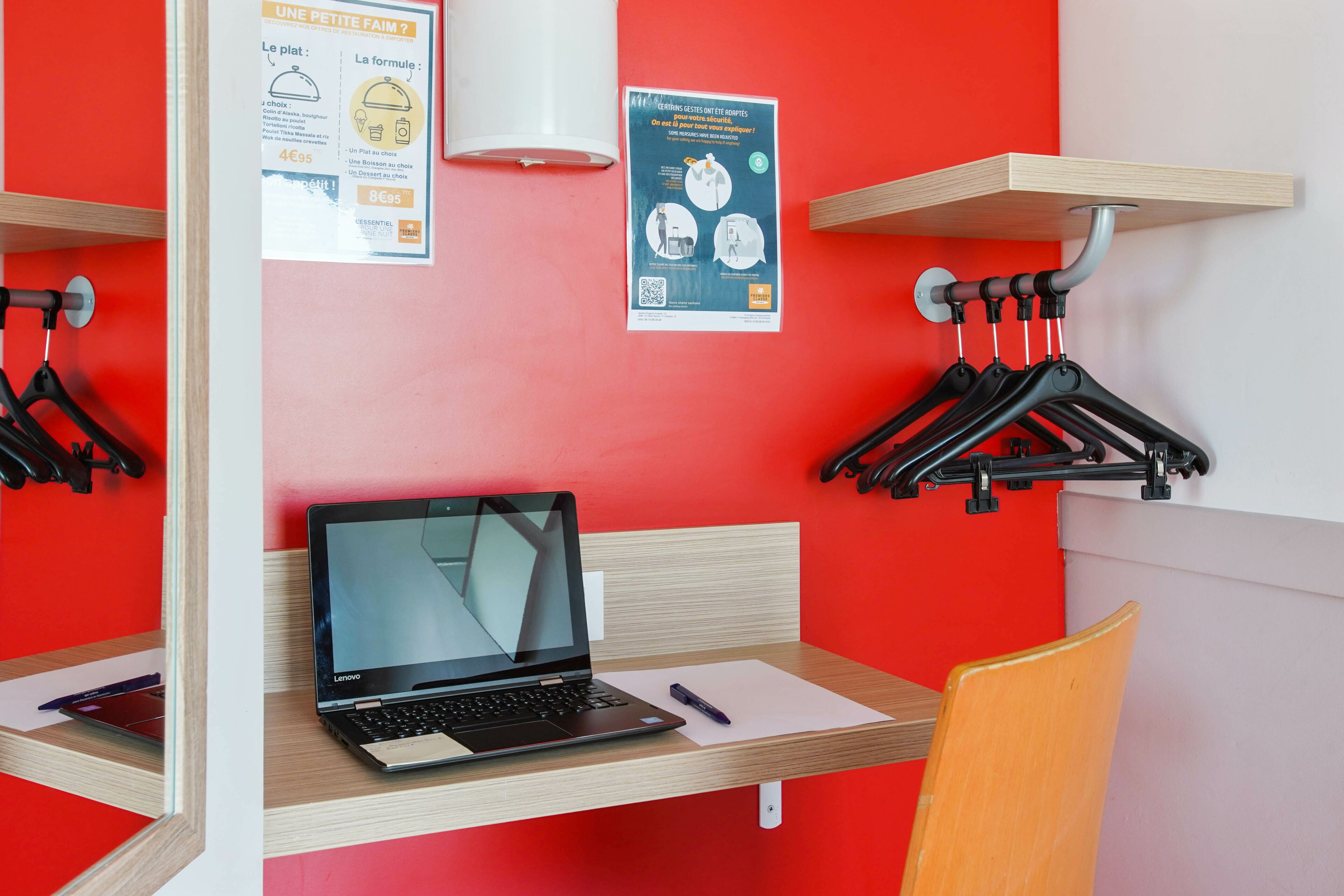 Un petit espace de travail comprenant un ordinateur portable, une chaise et un bureau. Il y a des affiches au mur et un porte-manteau avec des cintres.