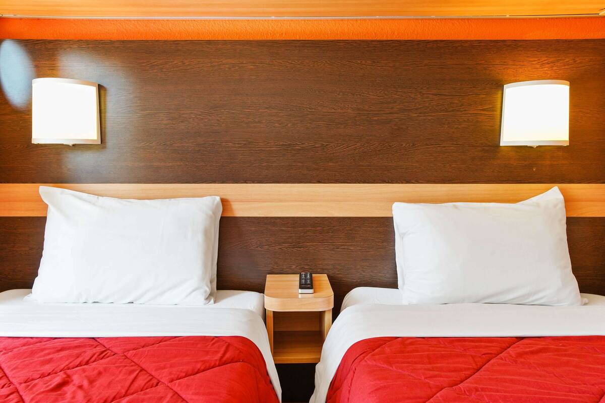 Une chambre d’hôtel avec deux lits, chacun doté de draps blancs et d’une couette rouge, d’une tête de lit en bois et d’une table de chevet avec une télécommande.