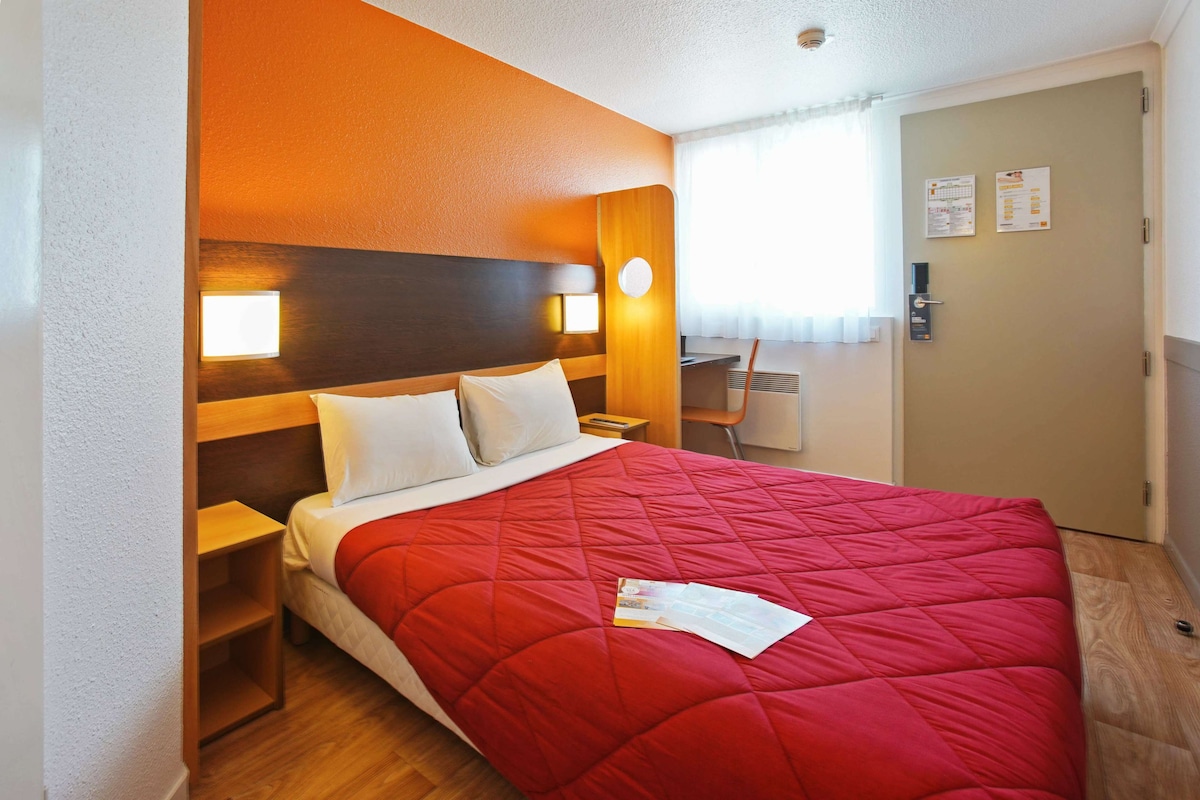 Een hotelkamer met een rode sprei, houten hoofdeinde, nachtkastjes en een bureau met een stoel.
