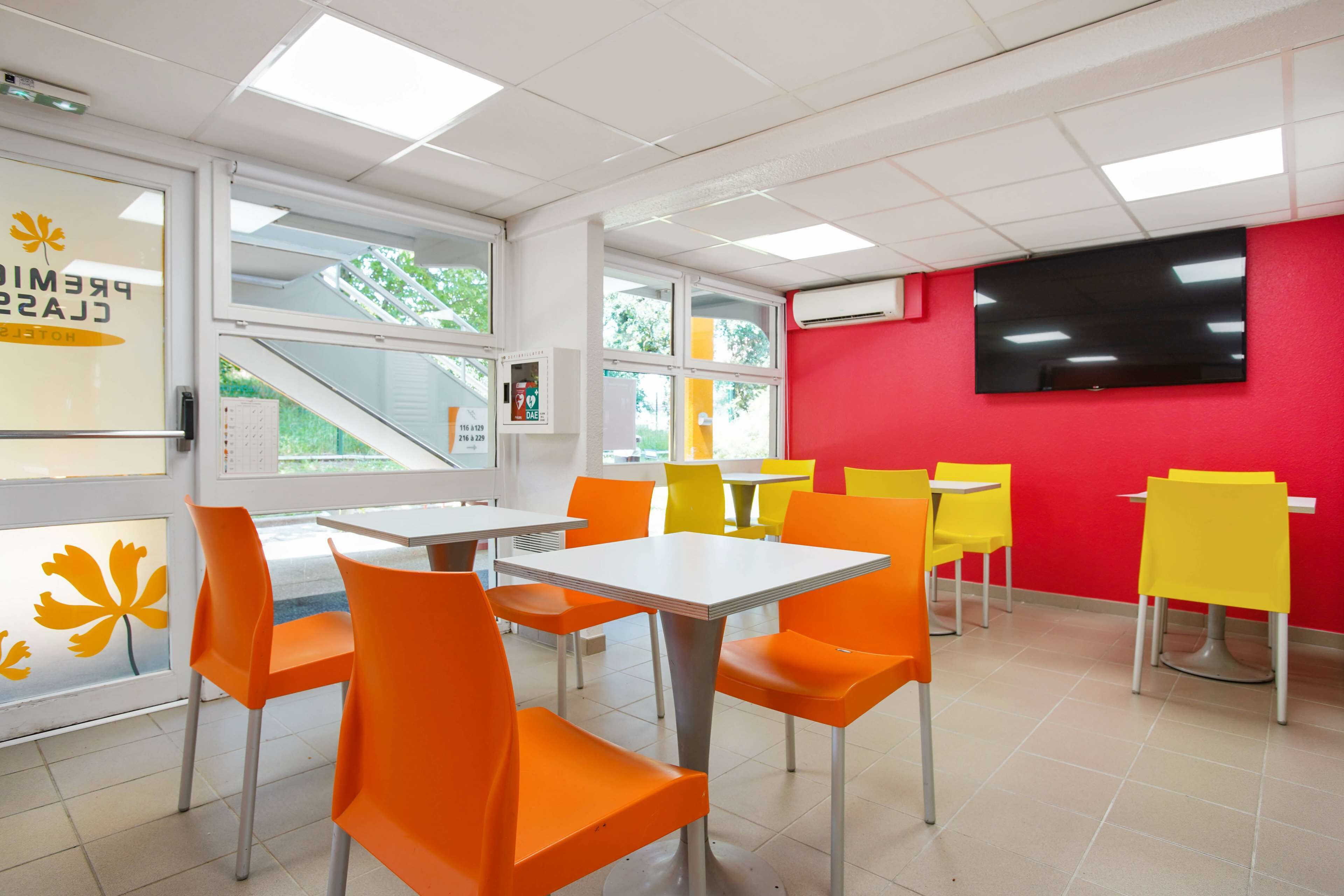 A brightly lit indoor space with orange and yellow chairs, white tables, and a red wall with a flat-screen TV.