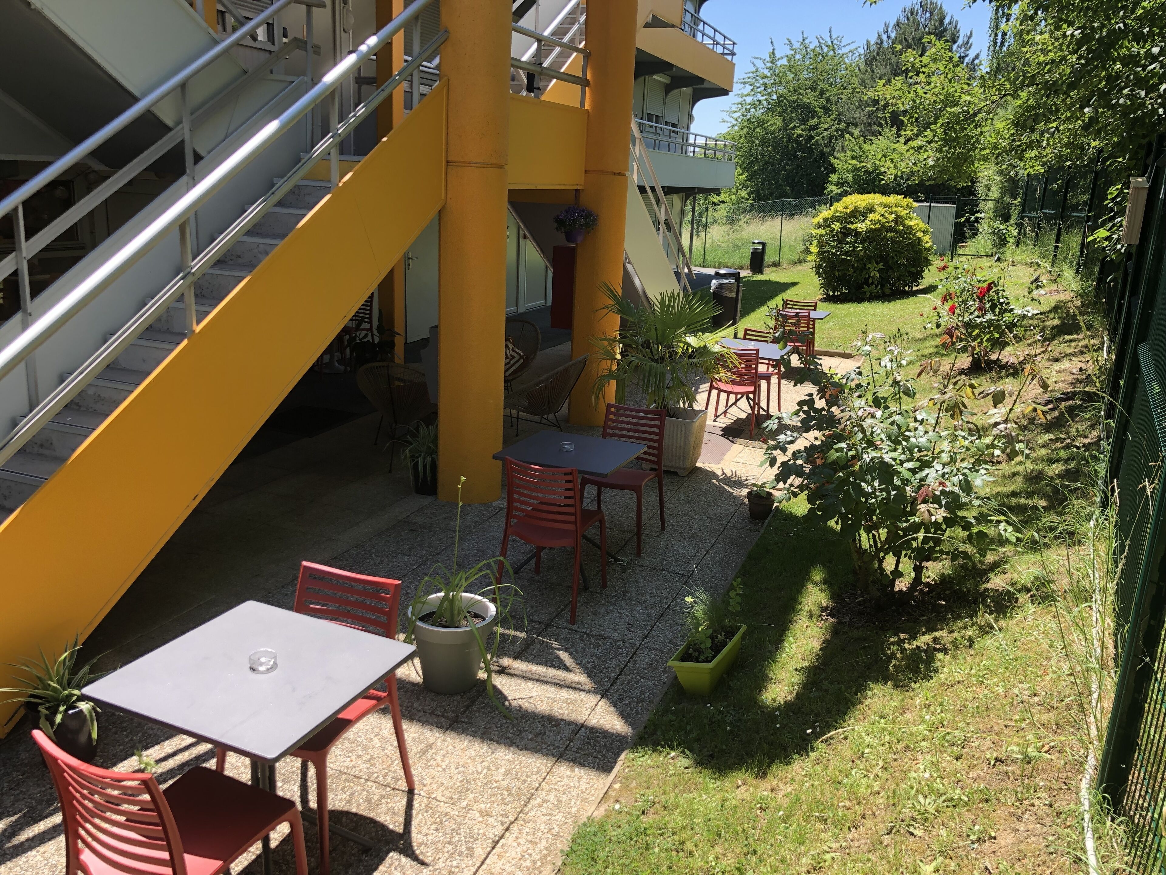An outdoor patio area with tables and chairs, surrounded by greenery and a yellow staircase.