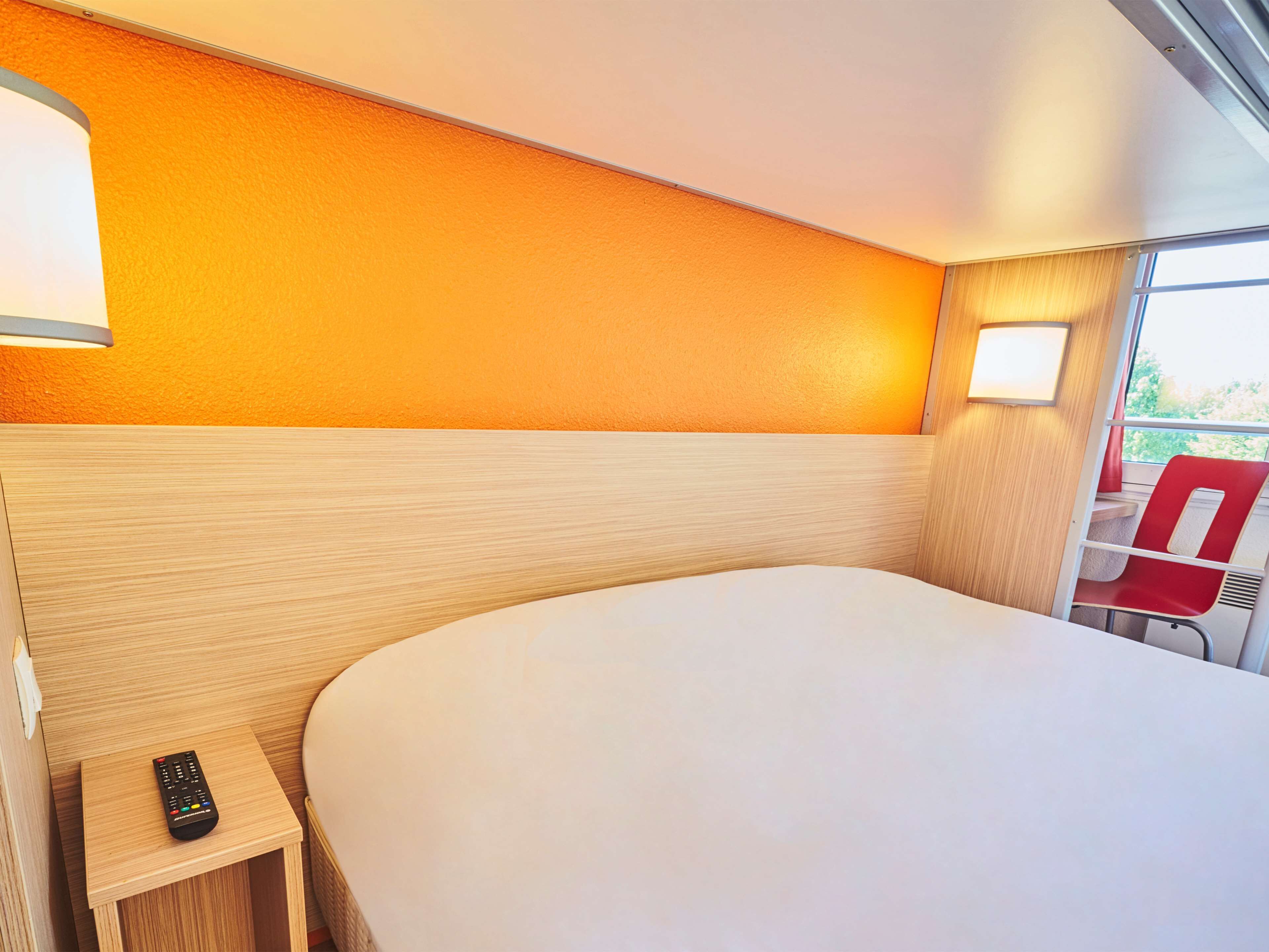Ein Hotelzimmer mit einem großen Bett, einem Nachttisch mit Fernbedienung und einem Fenster mit einem roten Sessel davor.