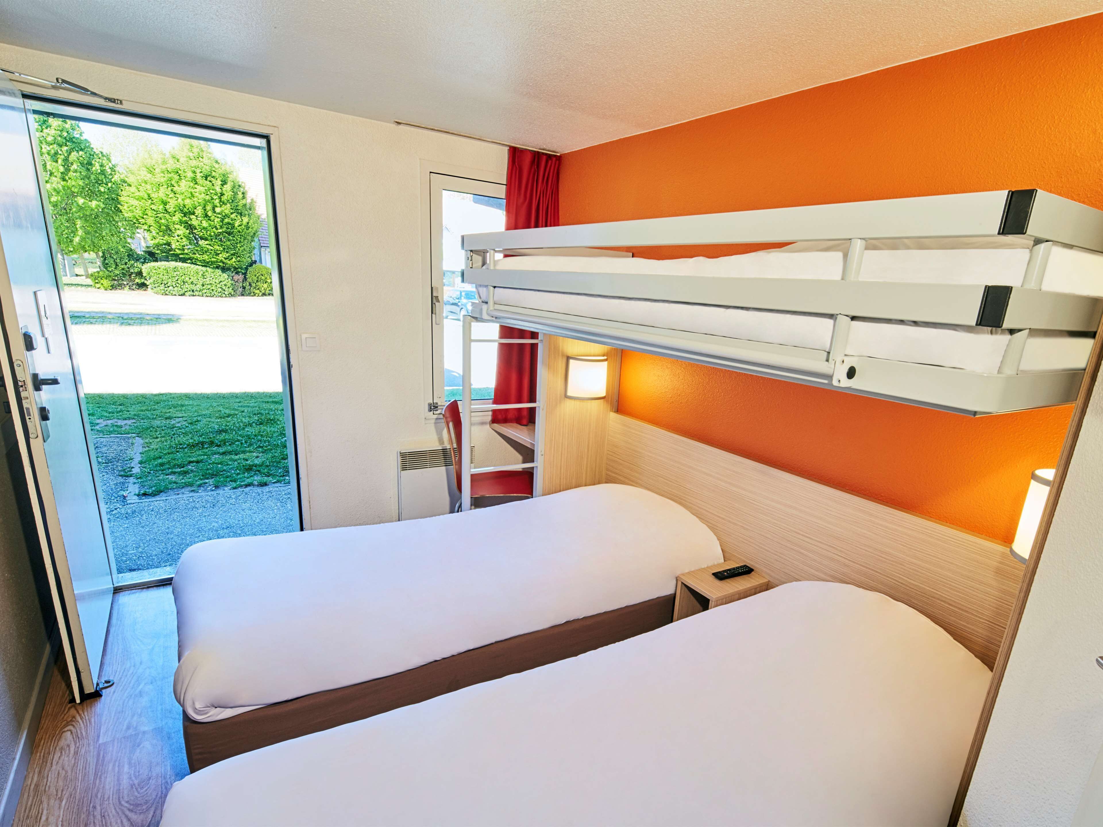 A compact hotel room with two beds, a small table, and a window with a view of greenery.