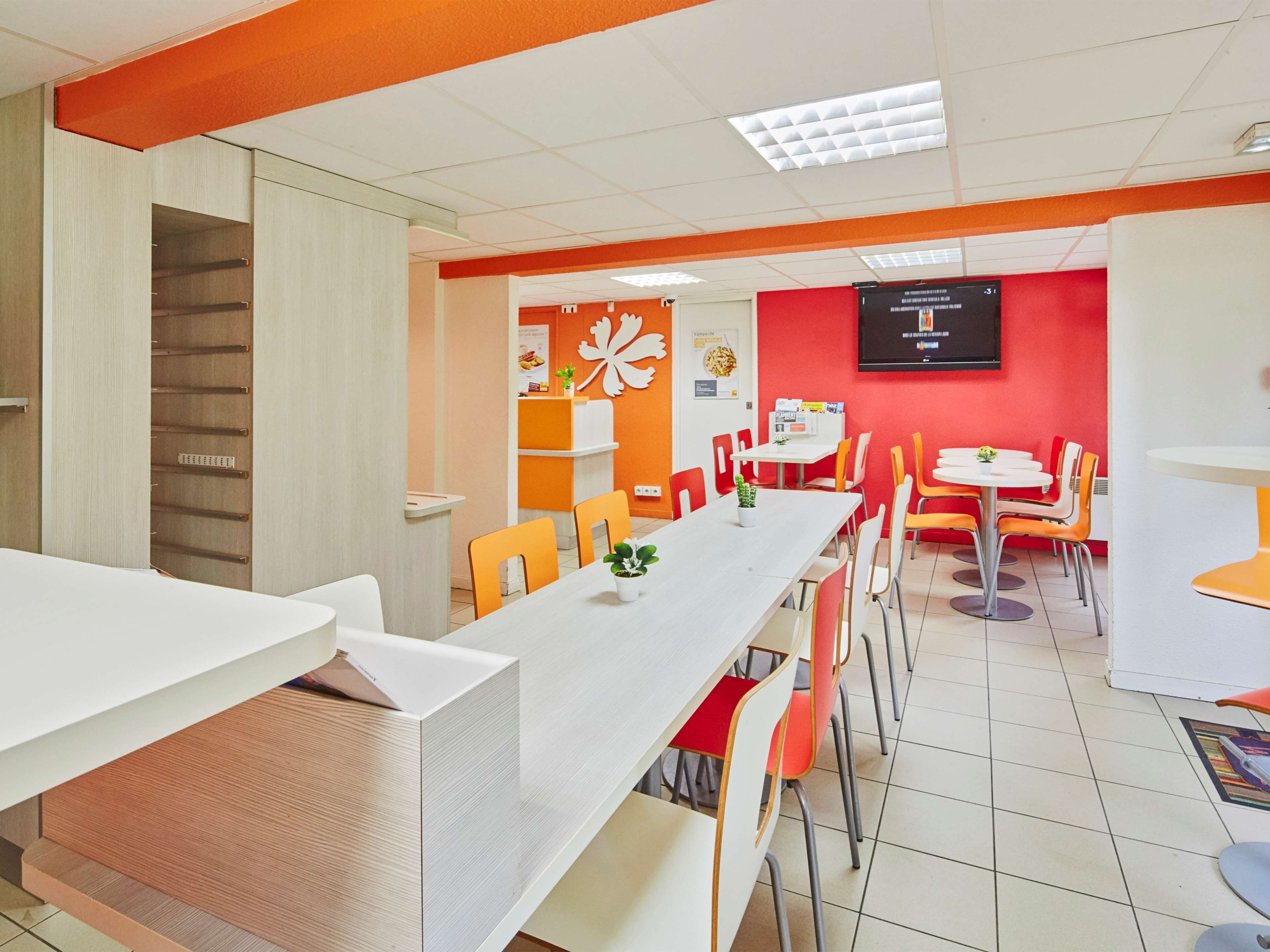 Un espace repas moderne avec des tables blanches et des chaises orange, un mur d’accent rouge et un panneau affichant le menu.
