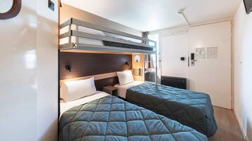 Ein Hotelzimmer mit zwei Betten, einem Holzkopfteil, einem kleinen Nachttisch, einer Wandleuchte und einer Tür mit Korkbrett.