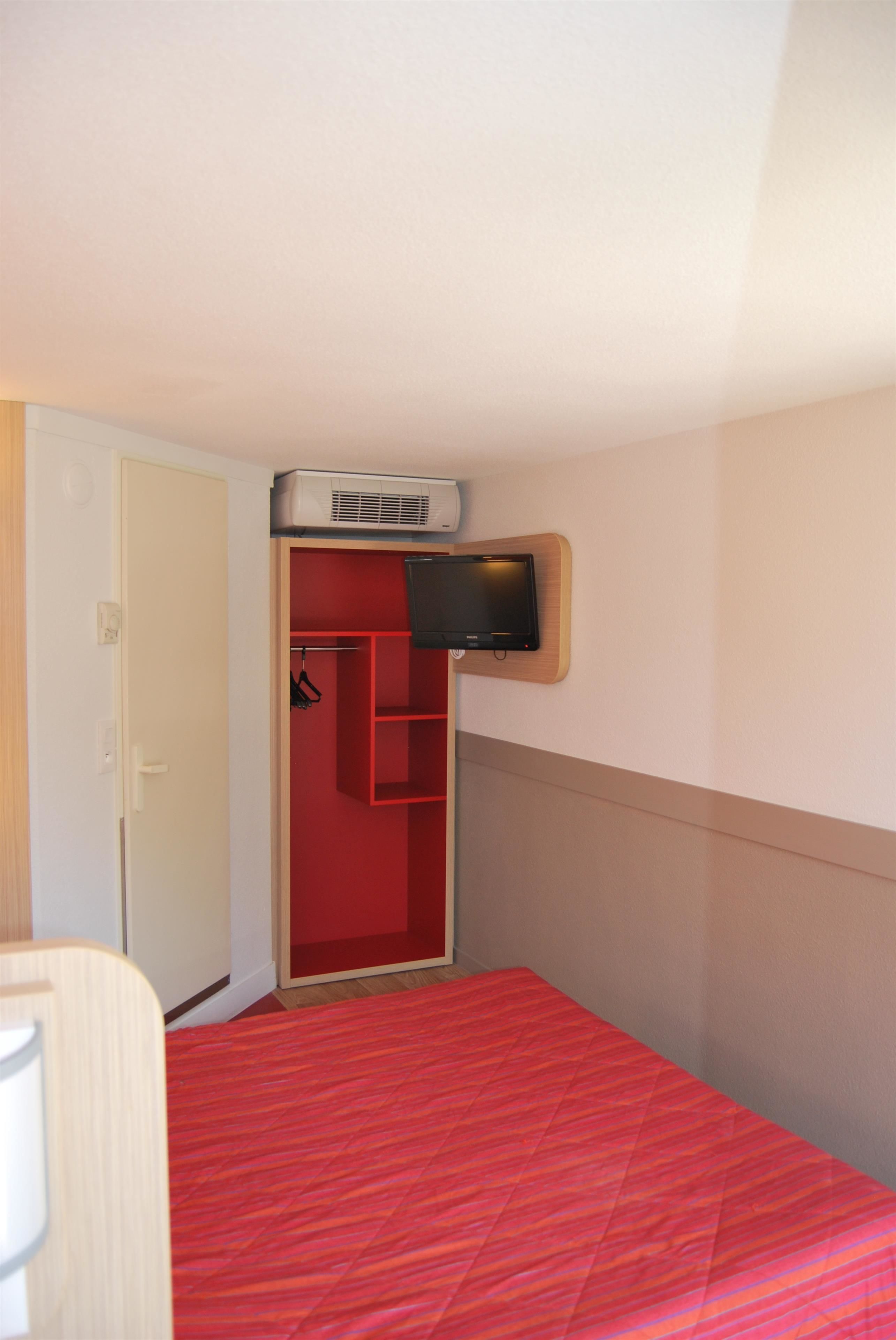 A room with a red bedspread, a television mounted on a wooden shelf, and a red wardrobe.