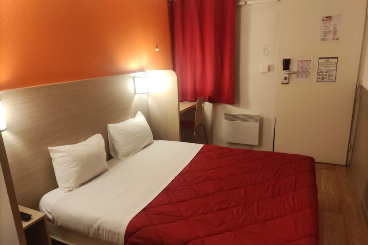 A hotel room with a bed, a desk, a chair, a radiator, and a door with a bulletin board.