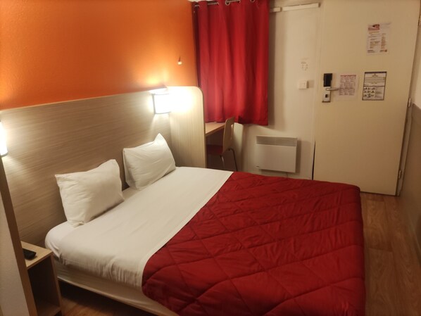 Habitación de hotel con cama, escritorio, silla, radiador y puerta con un tablón de anuncios.