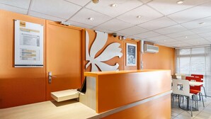 Un espace d’accueil comprenant un comptoir en bois, un panneau d’affichage avec un avis, et un coin salon avec des tables et des chaises.