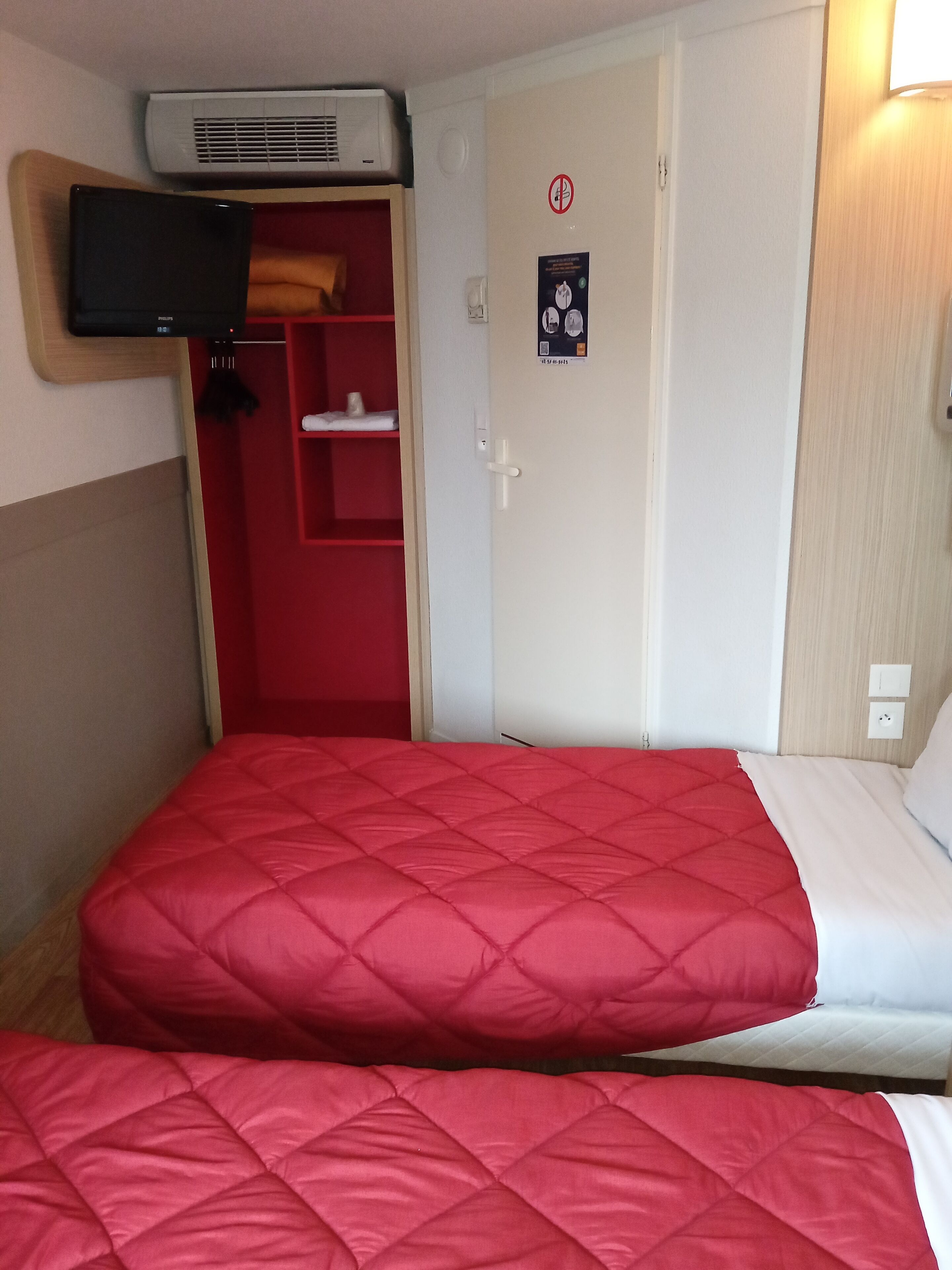 Une chambre avec deux lits, un téléviseur fixé au mur, un meuble de rangement rouge et une porte avec une pancarte.