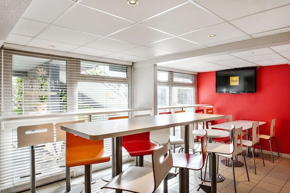 Un comedor moderno con varias mesas y sillas, ventanas amplias con persianas y un televisor de pantalla plana montado en una pared roja.
