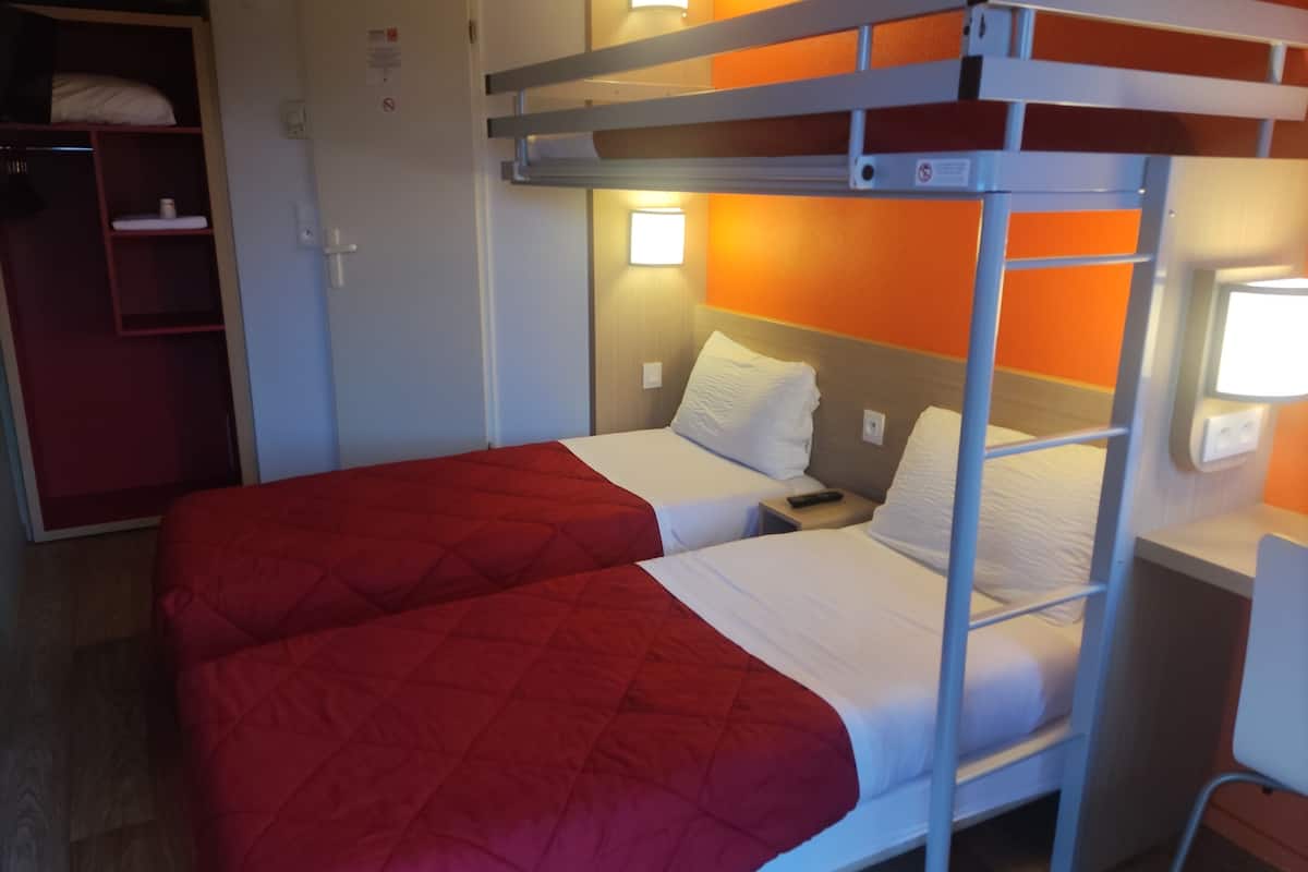 A bunk bed room with a red quilted bedspread, a wooden wardrobe, and a nightstand with a lamp.