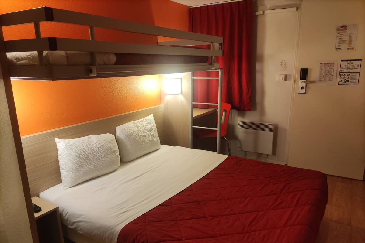 A bunk bed with a red quilt, a wooden headboard, and a bedside table with a remote control.