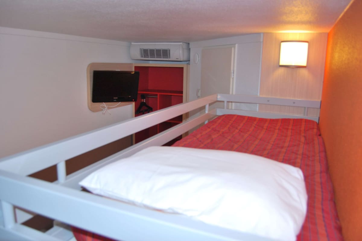 Una cama individual con colchón blanco y ropa de cama roja, cabecera blanca y un televisor fijado a la pared.
