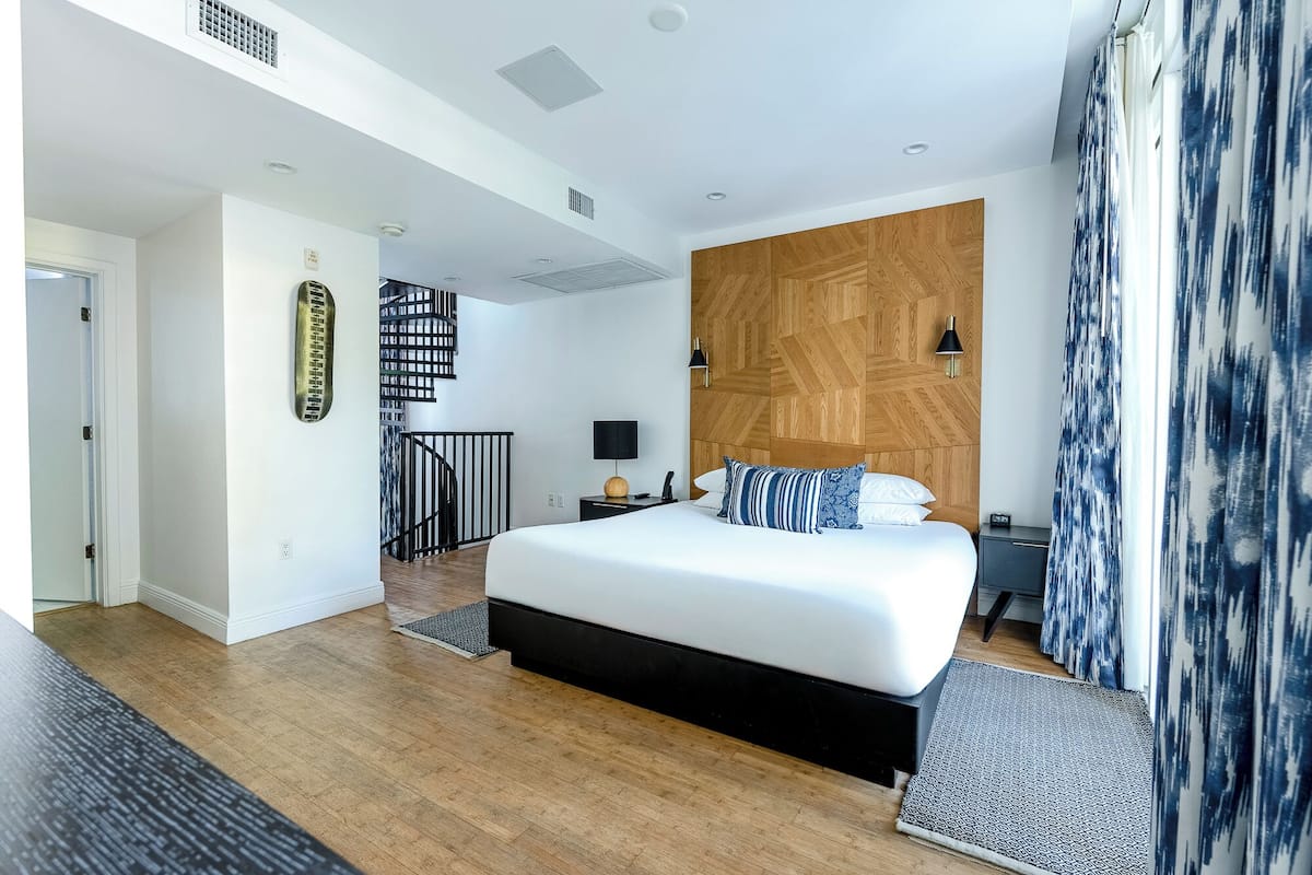 A modern hotel room with a large bed, a wooden headboard, a black nightstand, a wall-mounted TV, and a staircase leading to an upper level.
