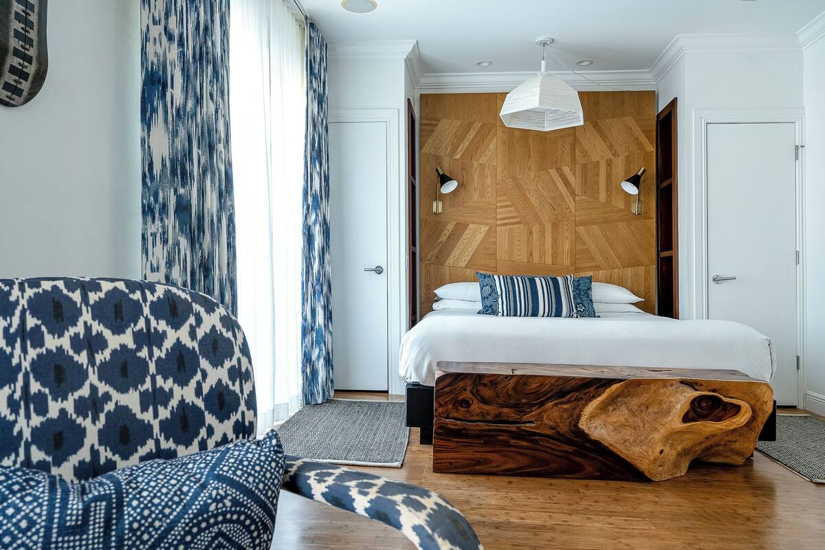 A modern hotel room with a large bed, a wooden headboard, a patterned armchair, and a unique wooden bench.