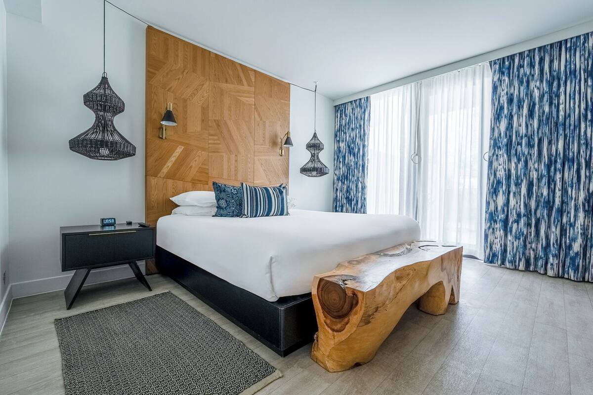A modern bedroom with a large bed, bedside tables, a wooden bench, and a large window with curtains.
