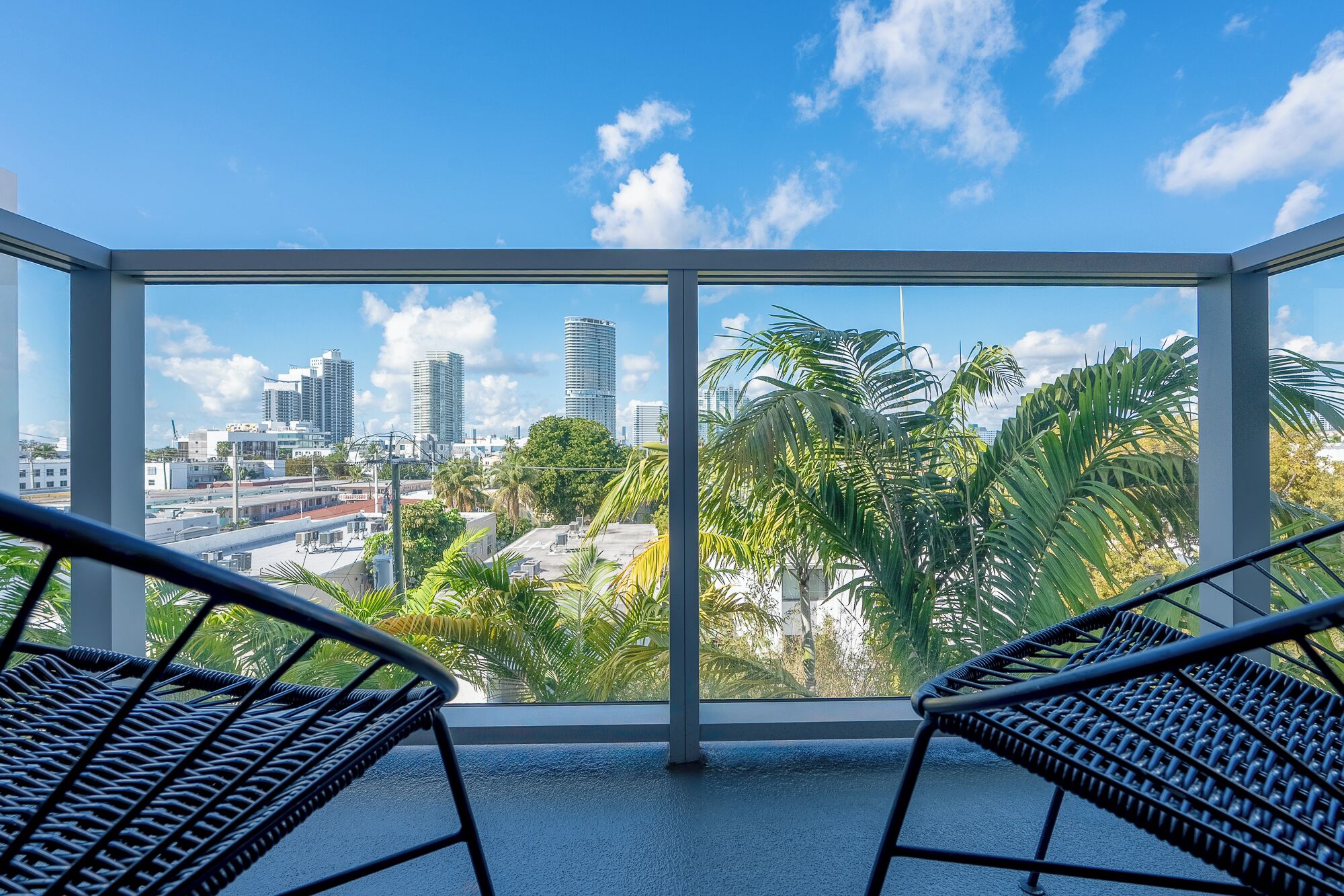 A balcony with two wicker chairs, a city view with palm trees, and tall buildings.