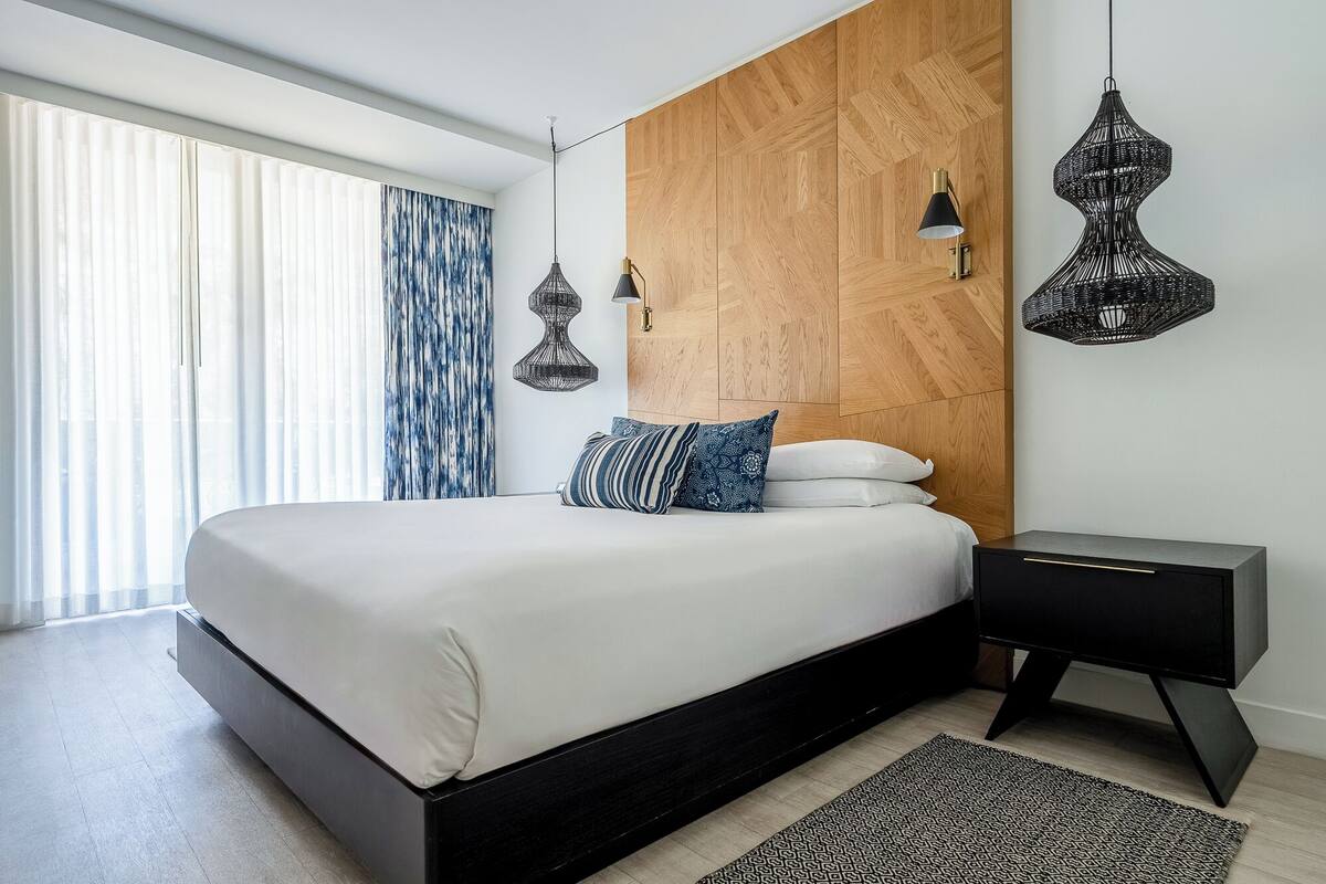 A modern bedroom with a large bed, a wooden headboard, two pendant lights, and a nightstand.