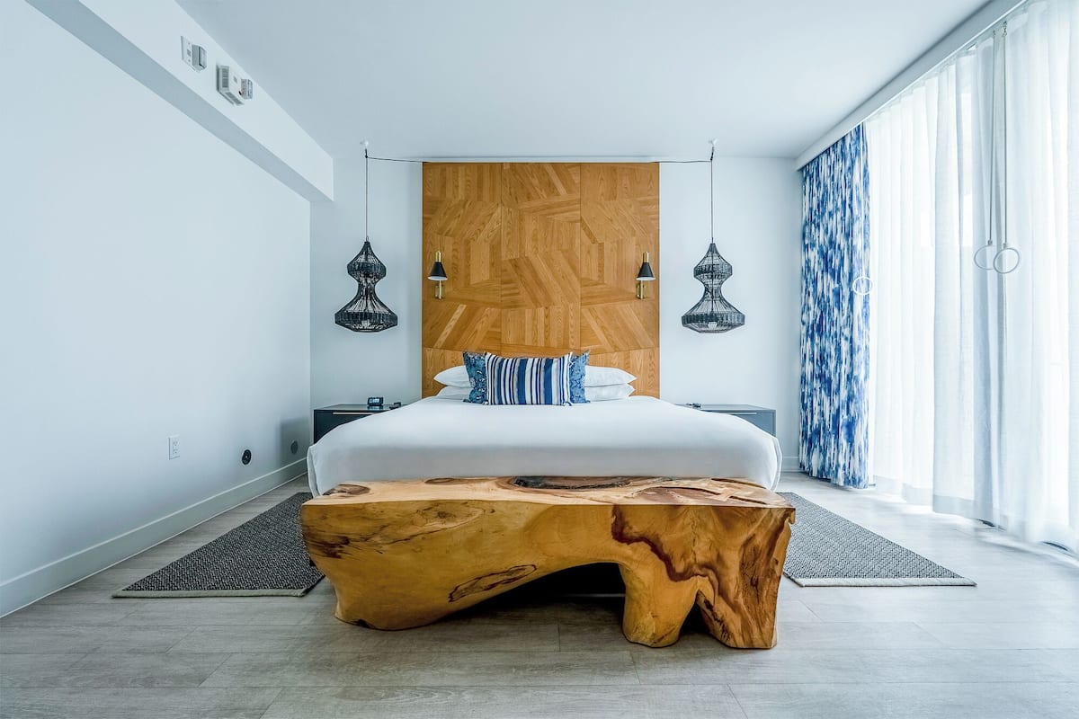 A modern bedroom with a large bed, a wooden headboard, two wall-mounted lamps, and a large window with curtains.