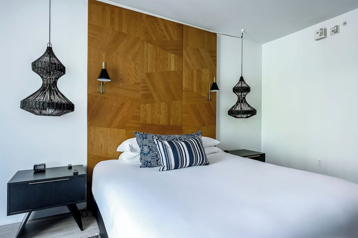 A modern bedroom with a wooden headboard, a bed with white bedding and patterned pillows, two black hanging lamps, and a small bedside table with a clock.