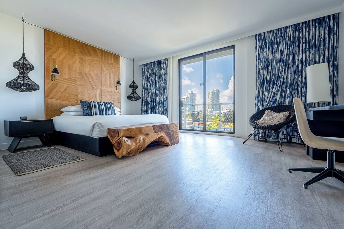 A modern bedroom with a large bed, a wooden headboard, a view of the city, and a balcony with chairs.