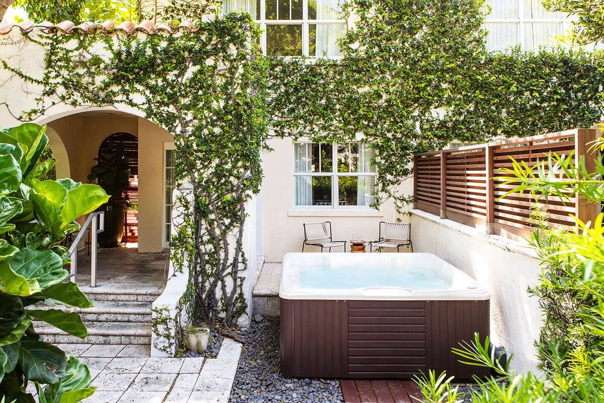 A hot tub in a garden with a wooden fence, a stone pathway, and a white building in the background.