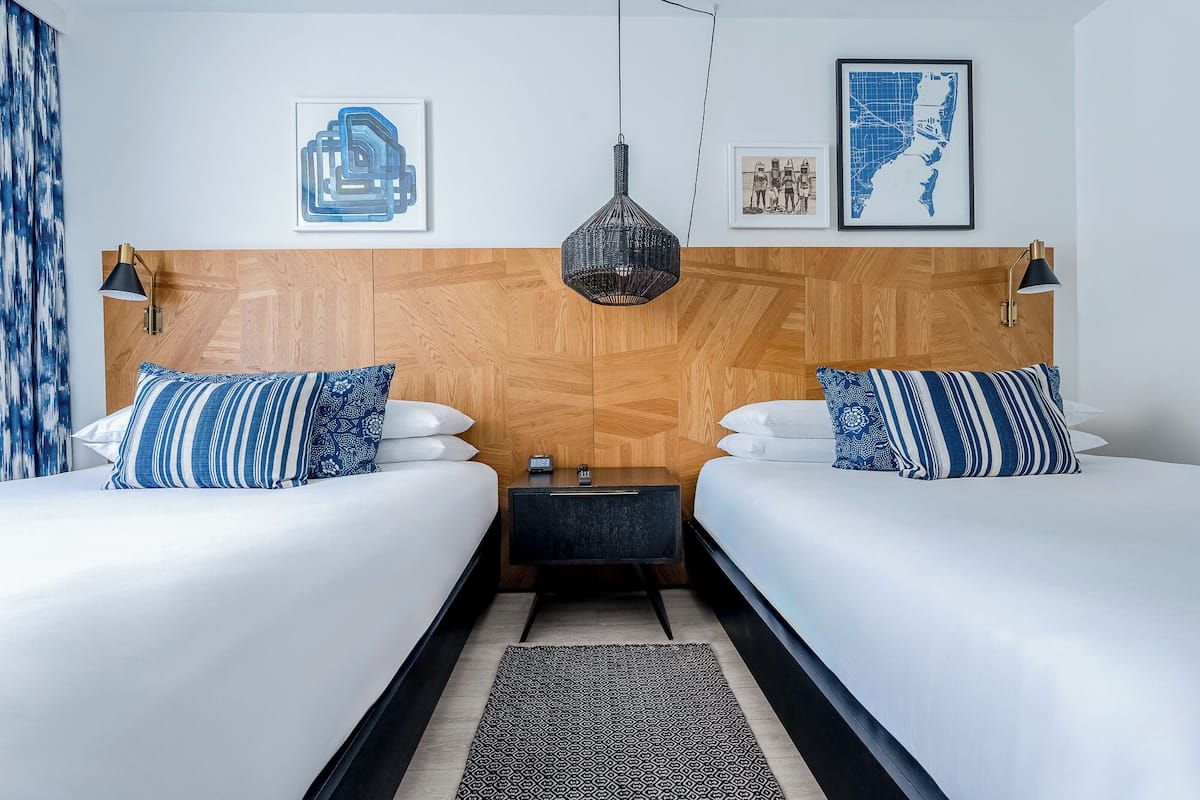 A hotel room with two beds, a wooden headboard, a black nightstand, a woven pendant light, and wall art.
