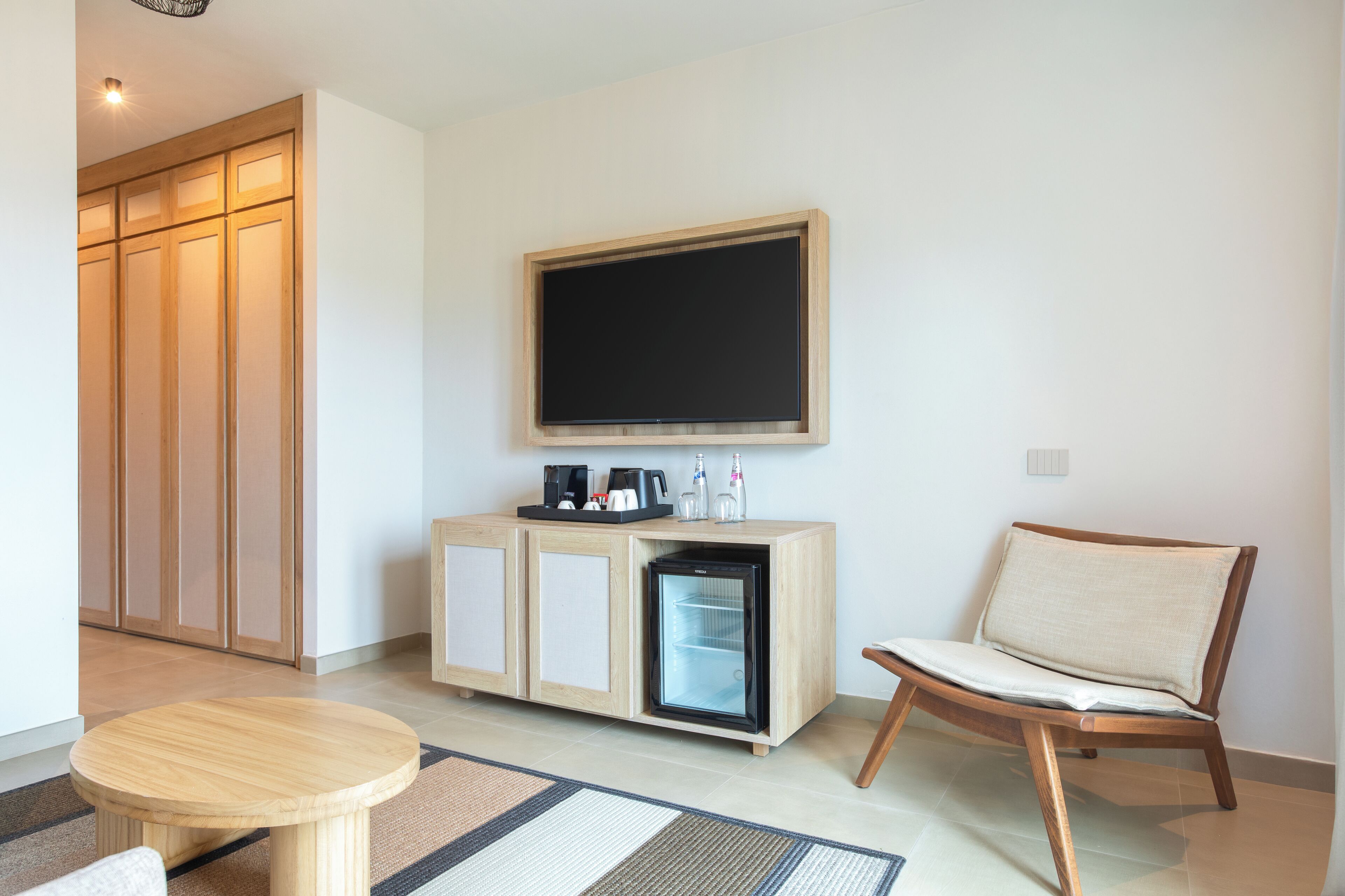 Una camera d'albergo con un armadietto in legno, un piccolo frigorifero, una sedia, un tavolino rotondo e una televisione a parete.