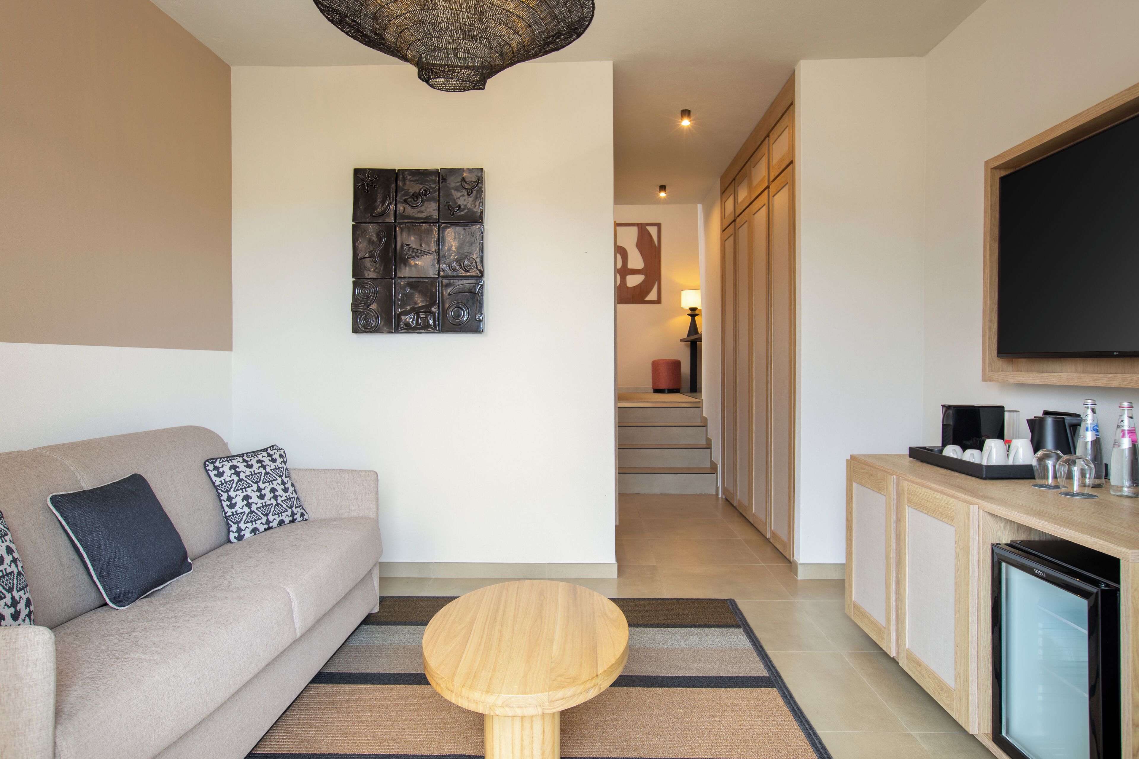 A modern living room with a sofa, a coffee table, a flat-screen TV, and a kitchen area with cabinets and a refrigerator.