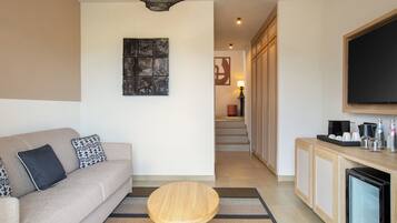 A modern living room with a sofa, a coffee table, a flat-screen TV, and a kitchen area with cabinets and a refrigerator.