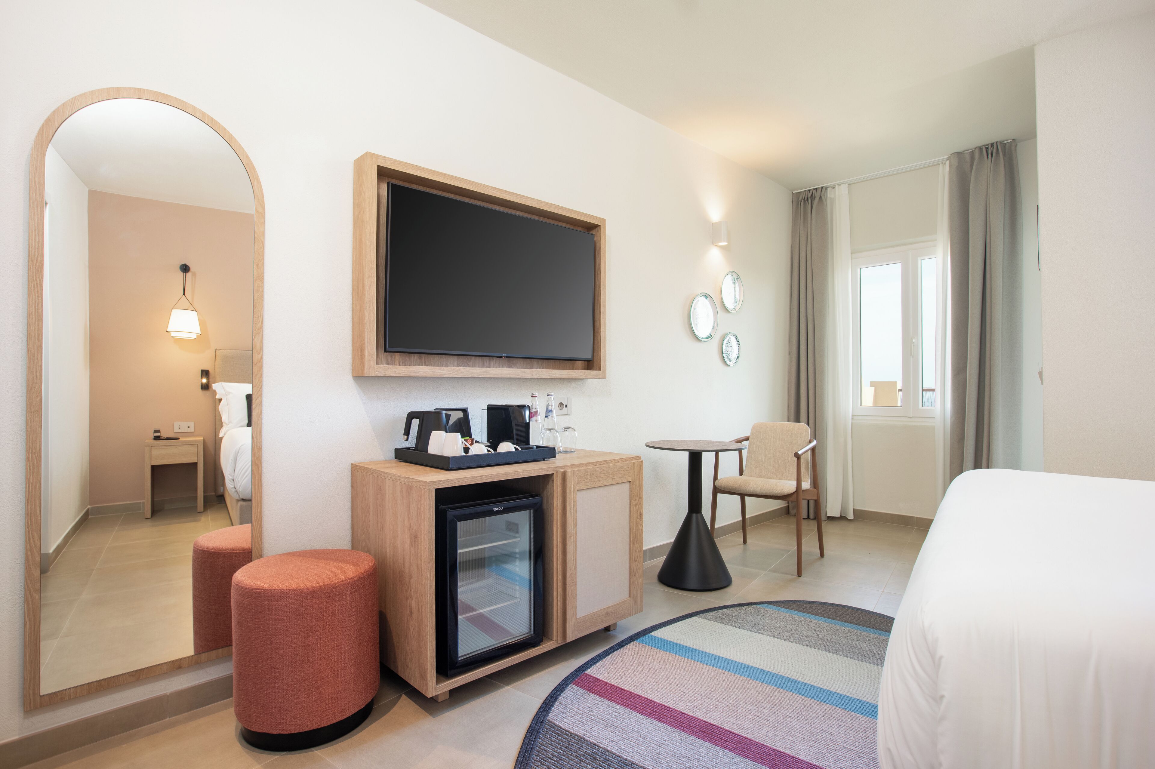 A hotel room with a bed, a small refrigerator, a coffee machine, a desk, and a chair.