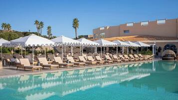 A poolside area with lounge chairs and umbrellas, a clear pool, and a building with a tiled roof in the background.