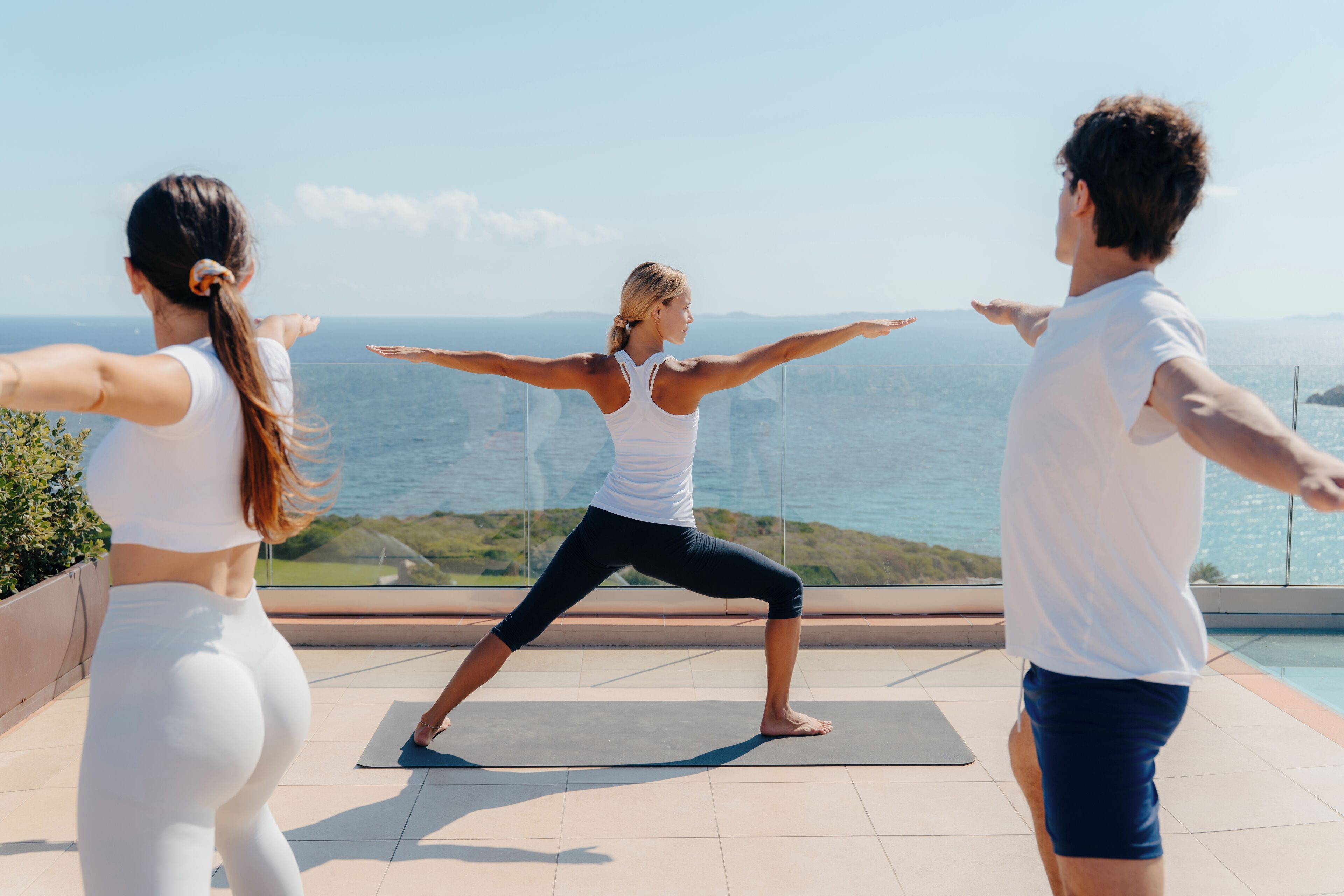 Trois personnes pratiquent le yoga sur un tapis, avec une vue panoramique sur l’océan.