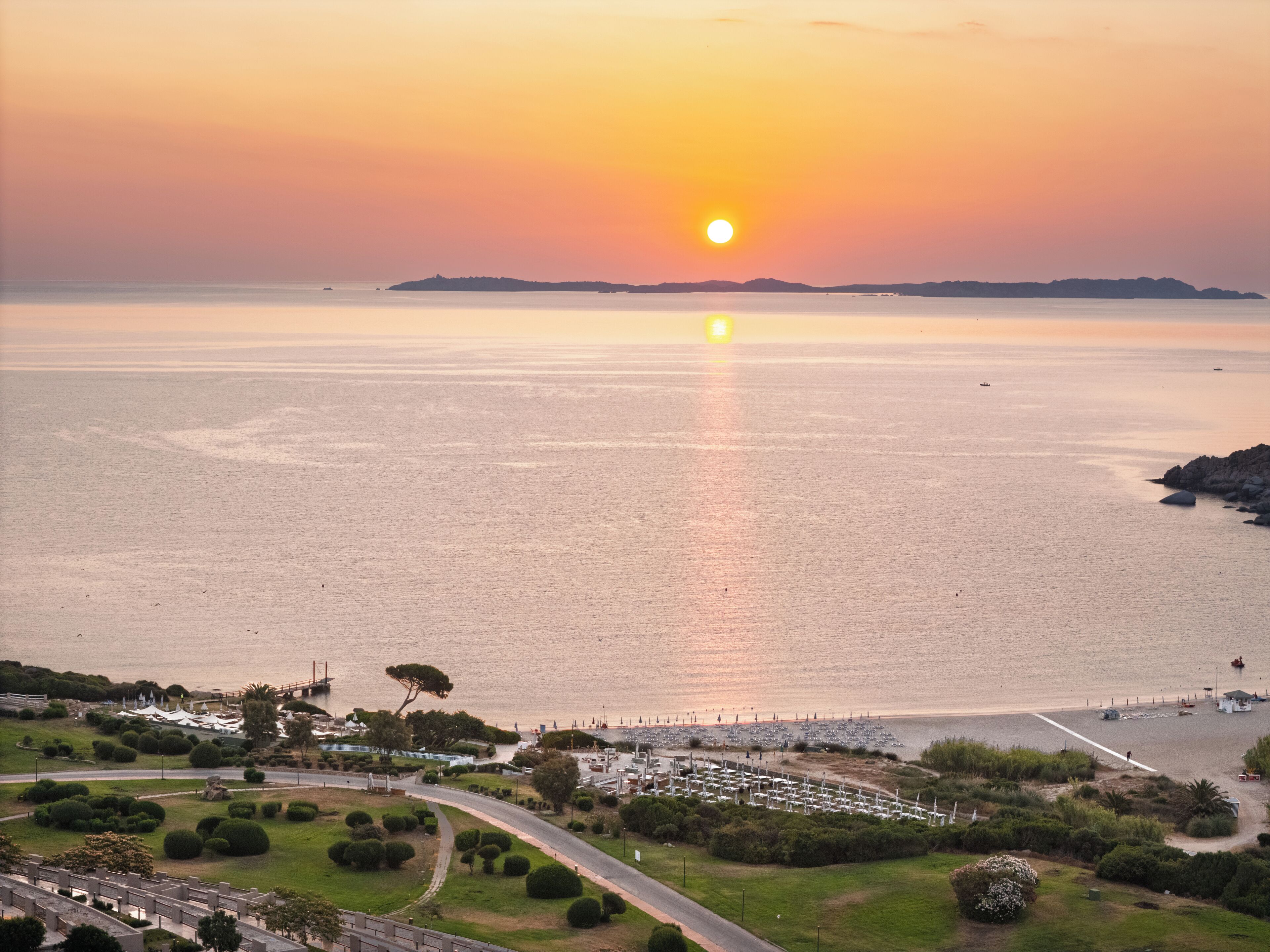 A coastal landscape with a sunset over the sea, a marina with boats, and a park with pathways and greenery.