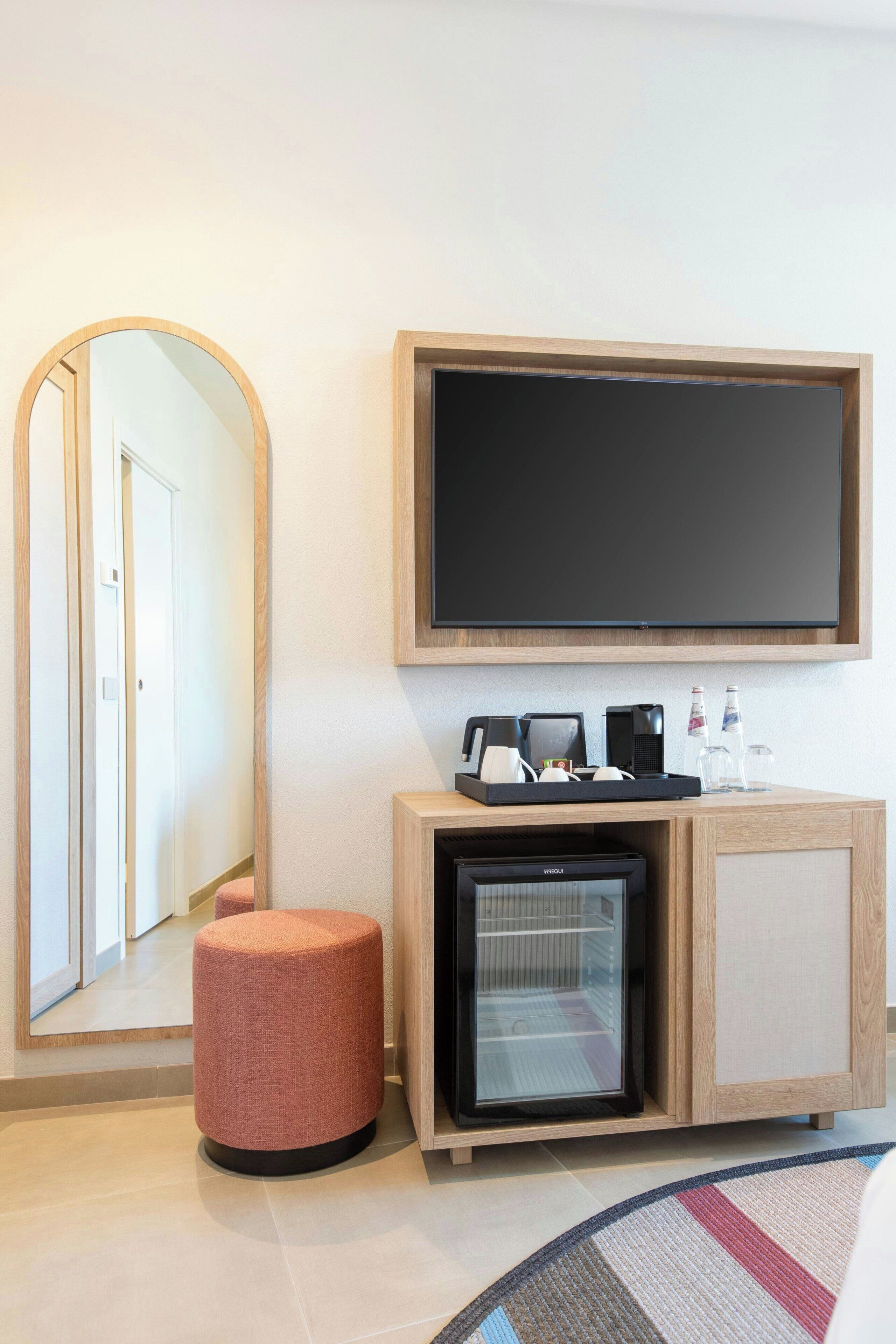 Hotellihuone, jossa on suuri peili, taulutelevisio, minibaari, jääkaappi ja pieni jalkarahi.