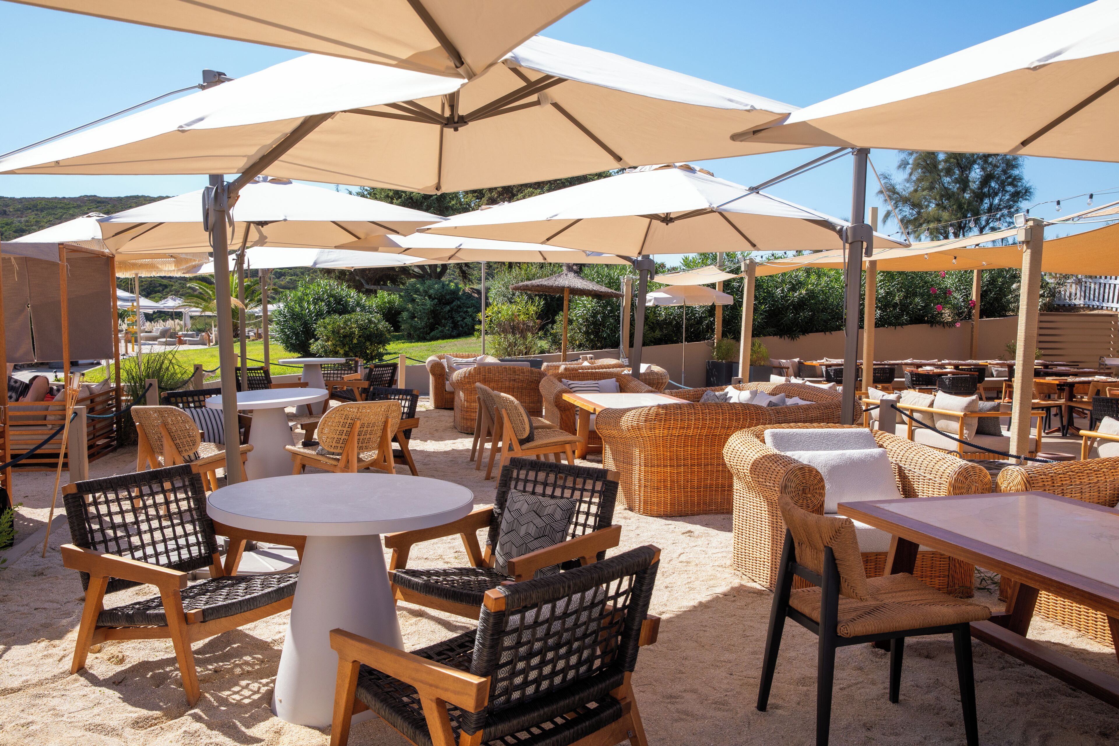 An outdoor seating area with round tables and wicker chairs, shaded by large umbrellas.