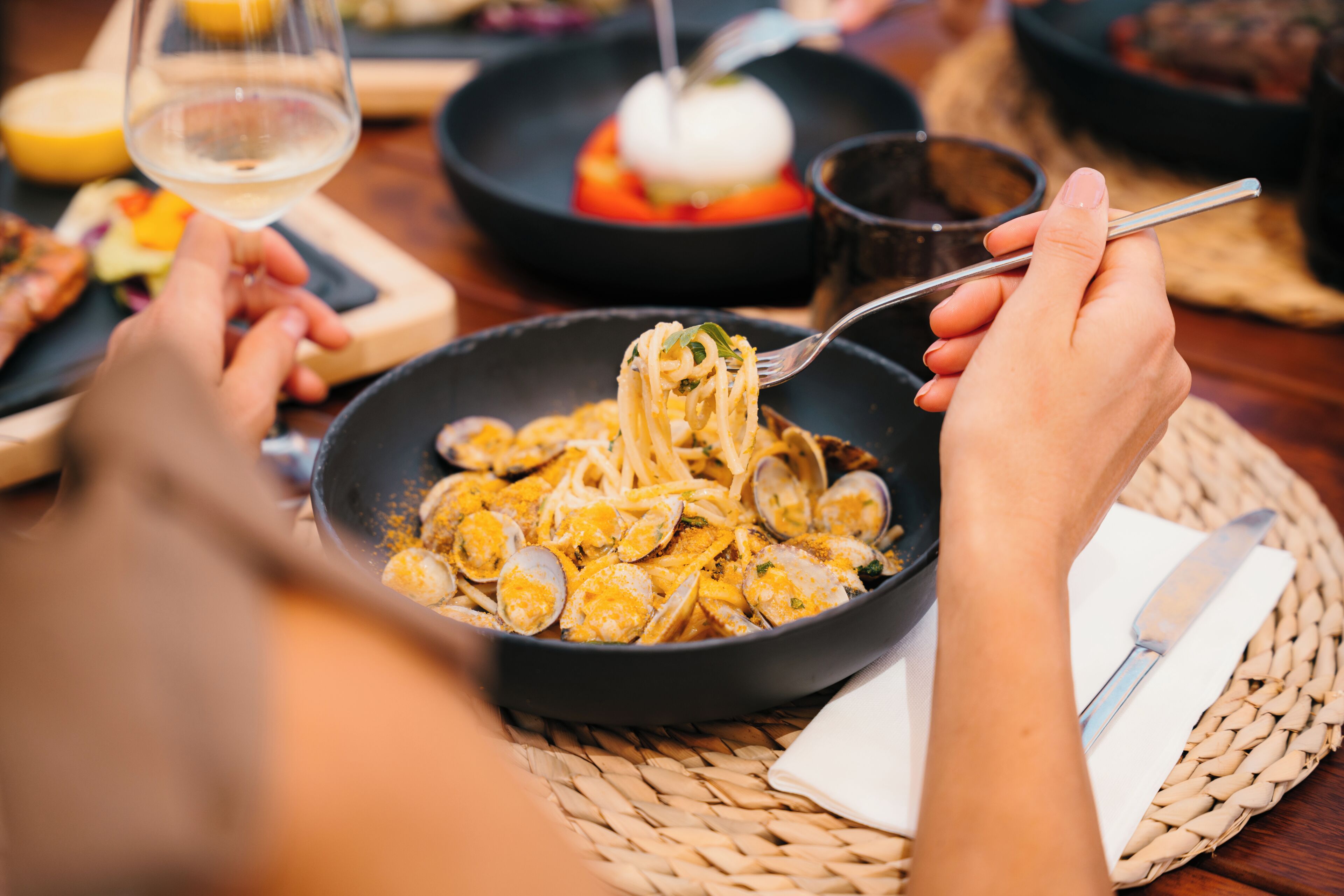 A dining table with a plate of pasta and clams, a fork, and a glass of white wine.