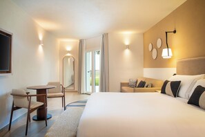 Een hotelkamer met een groot bed, een bank, een kleine tafel en een stoel. Er is een raam met gordijnen, een wandlamp en een spiegel.