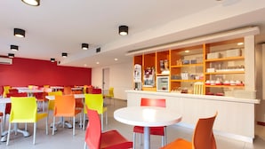 A brightly lit cafeteria with red and yellow chairs, a white counter with various food items, and overhead lighting. - Première Classe Dunkerque - Saint-Pol-Sur-Mer (Dunkirk)