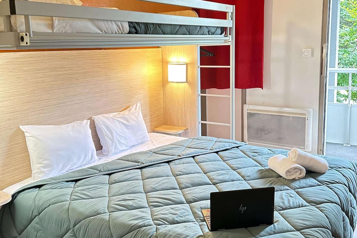 A bunk bed with a laptop on the lower bunk, a bedside table, and a window with red curtains.