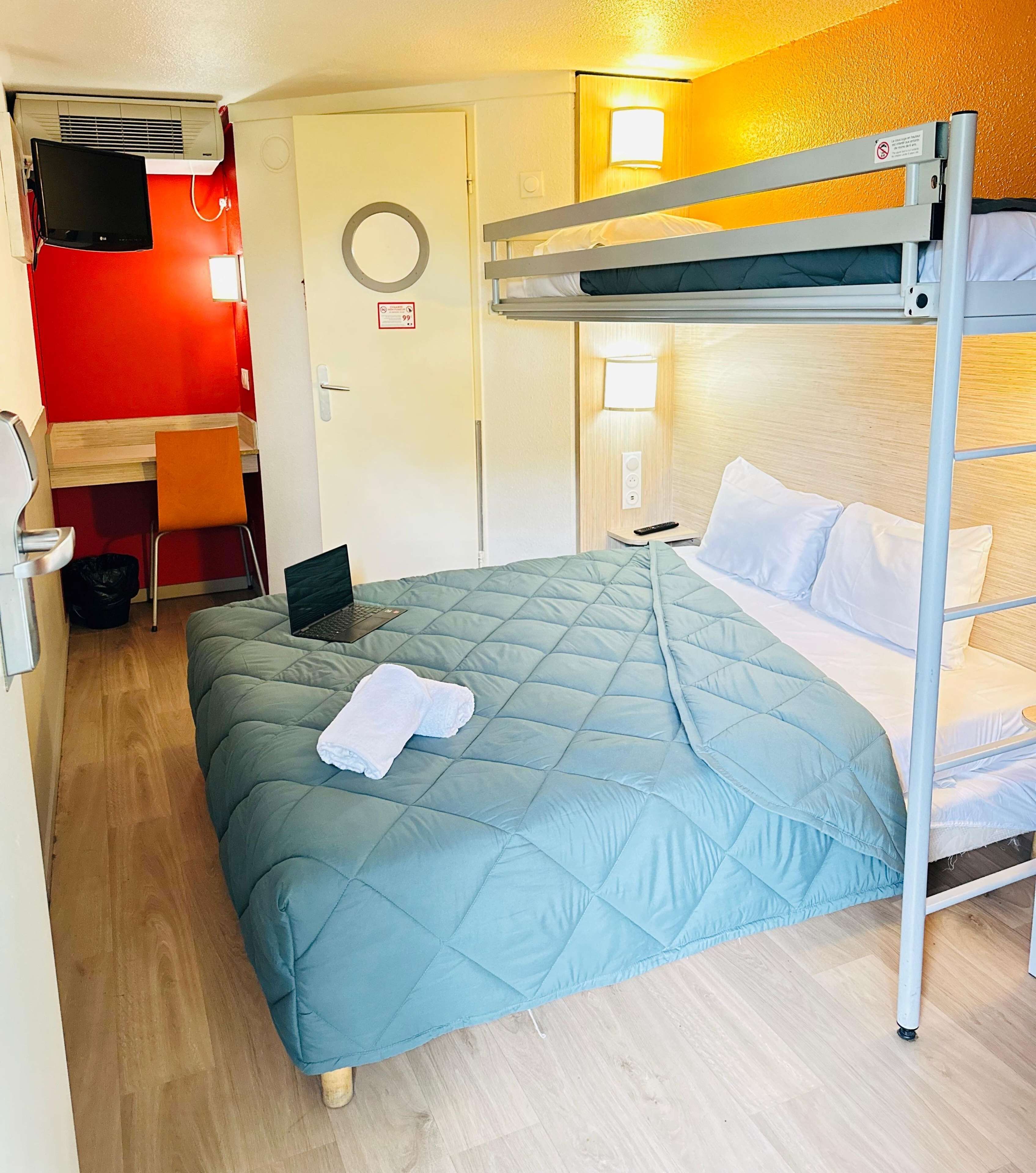 A bunk bed with a blue quilt, a desk with a laptop, a chair, and a red wall.
