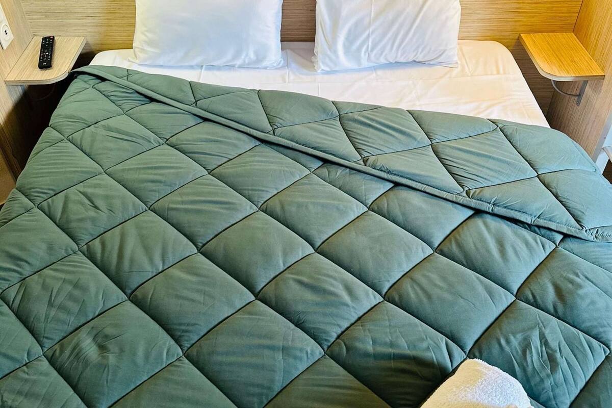A neatly made bed with a green quilt, white pillows, and a remote control on a wooden nightstand.