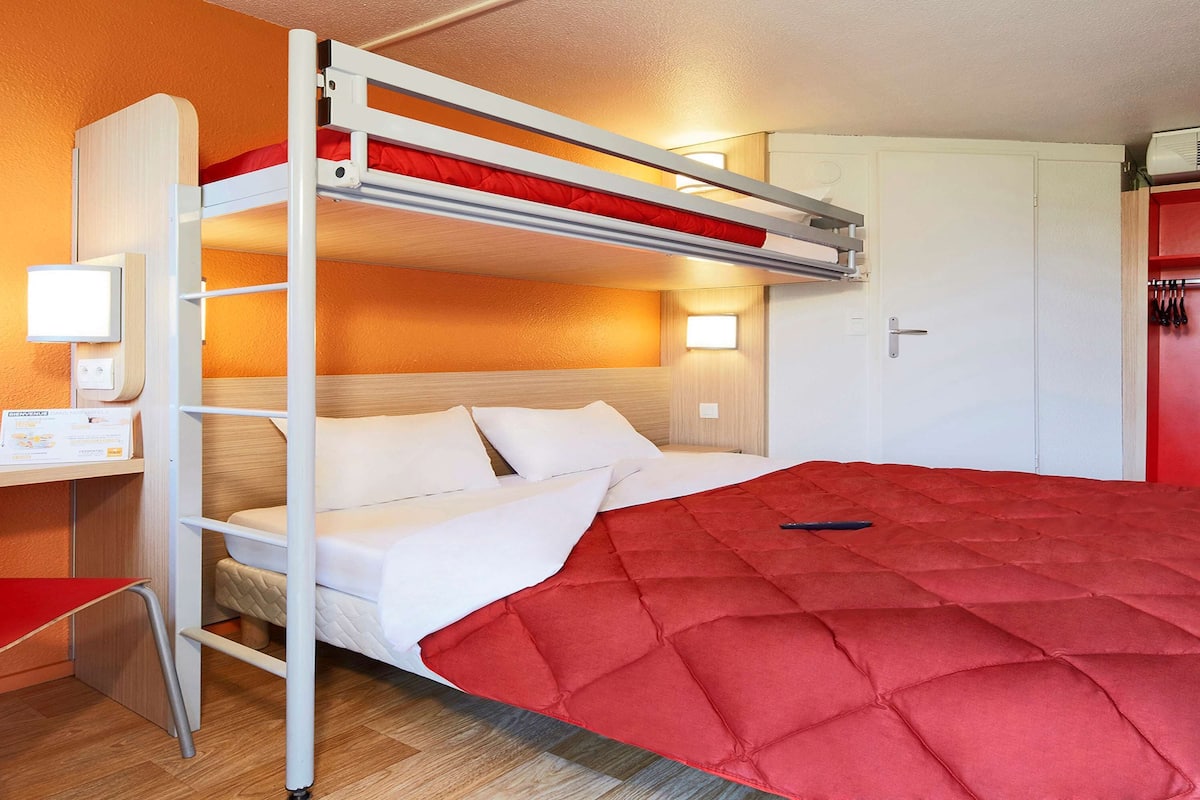 A bunk bed with a red mattress, a desk with a chair, a red wardrobe, and a wall-mounted TV.