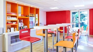 Une cafétéria bien éclairée, avec des meubles aux couleurs orange et blanc, un comptoir où sont exposés divers produits, et une grande fenêtre donnant sur un paysage verdoyant.