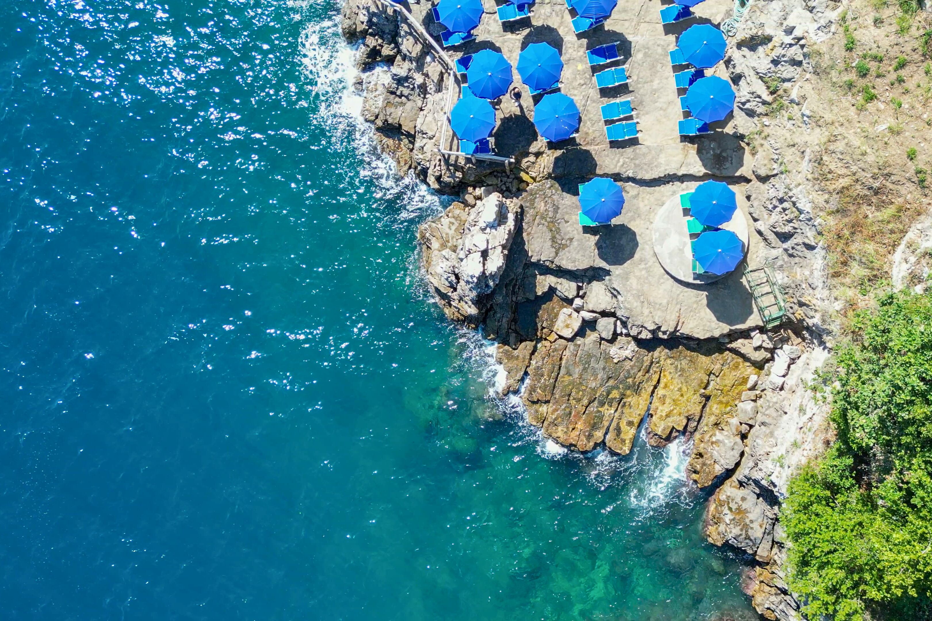 An aerial view of a coastal area with a rocky platform, clear blue water, and several blue umbrellas.