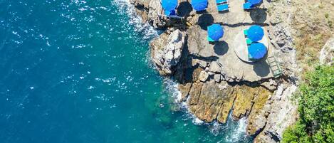 An aerial view of a coastal area with a rocky platform, clear blue water, and several blue umbrellas.