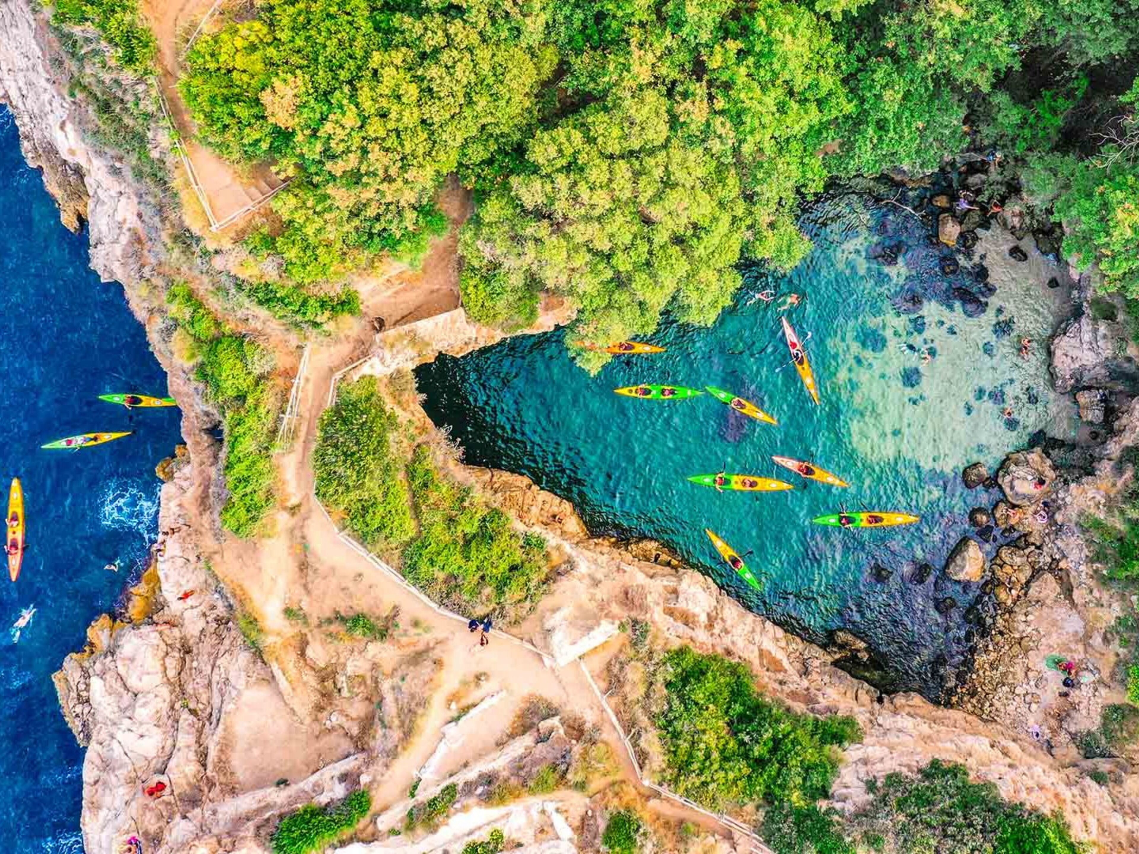Vue aérienne d’une zone côtière, avec des kayaks sur l’eau, entourée de falaises et de végétation.