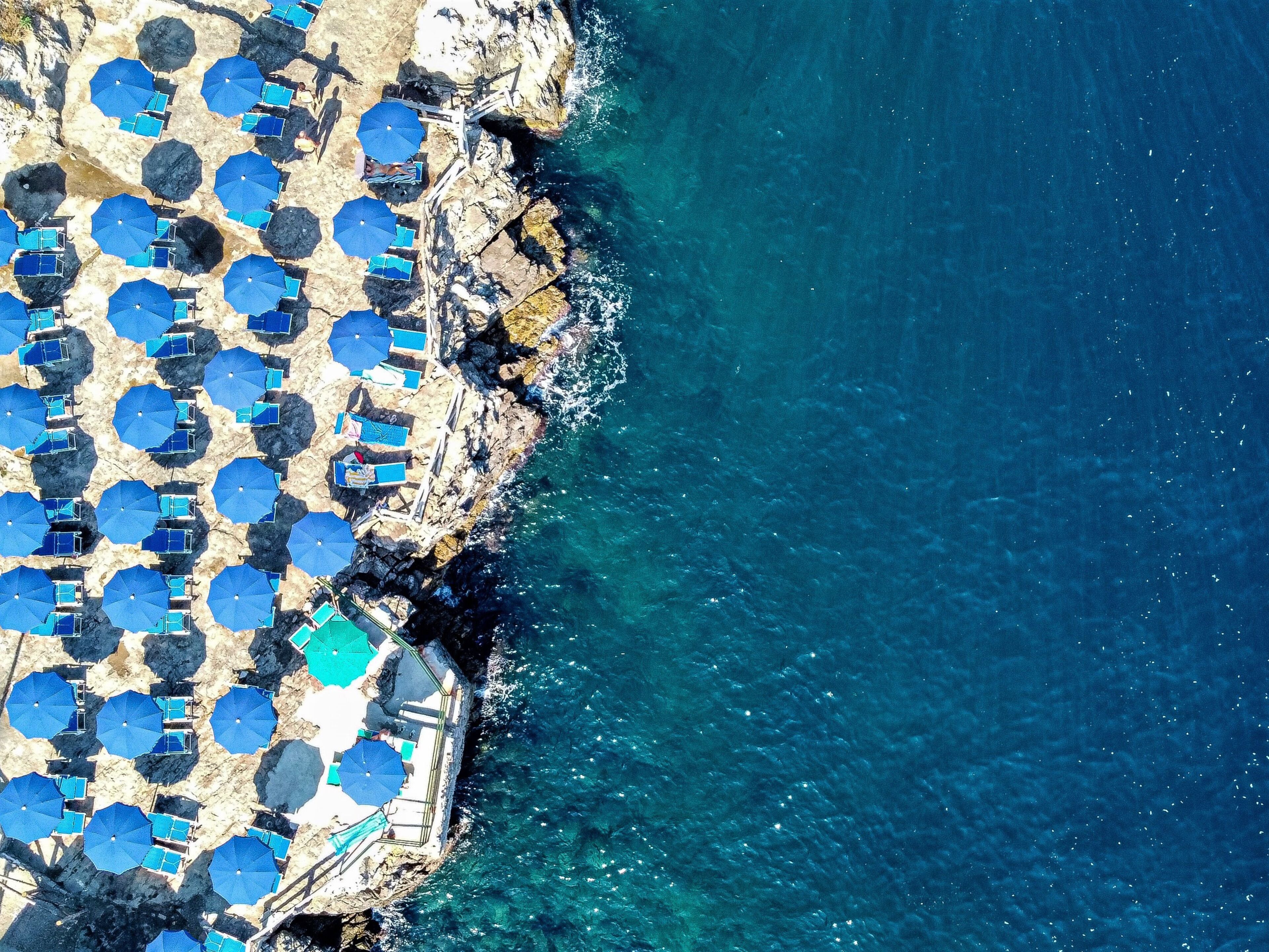 An aerial view of a coastal area with numerous blue umbrellas and sun loungers arranged along the shoreline, suggesting a beach resort setting.
